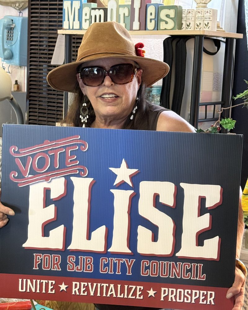 A supporter in a wide-brim hat holding a Vote Elise for SJB City Council sign inside a shop